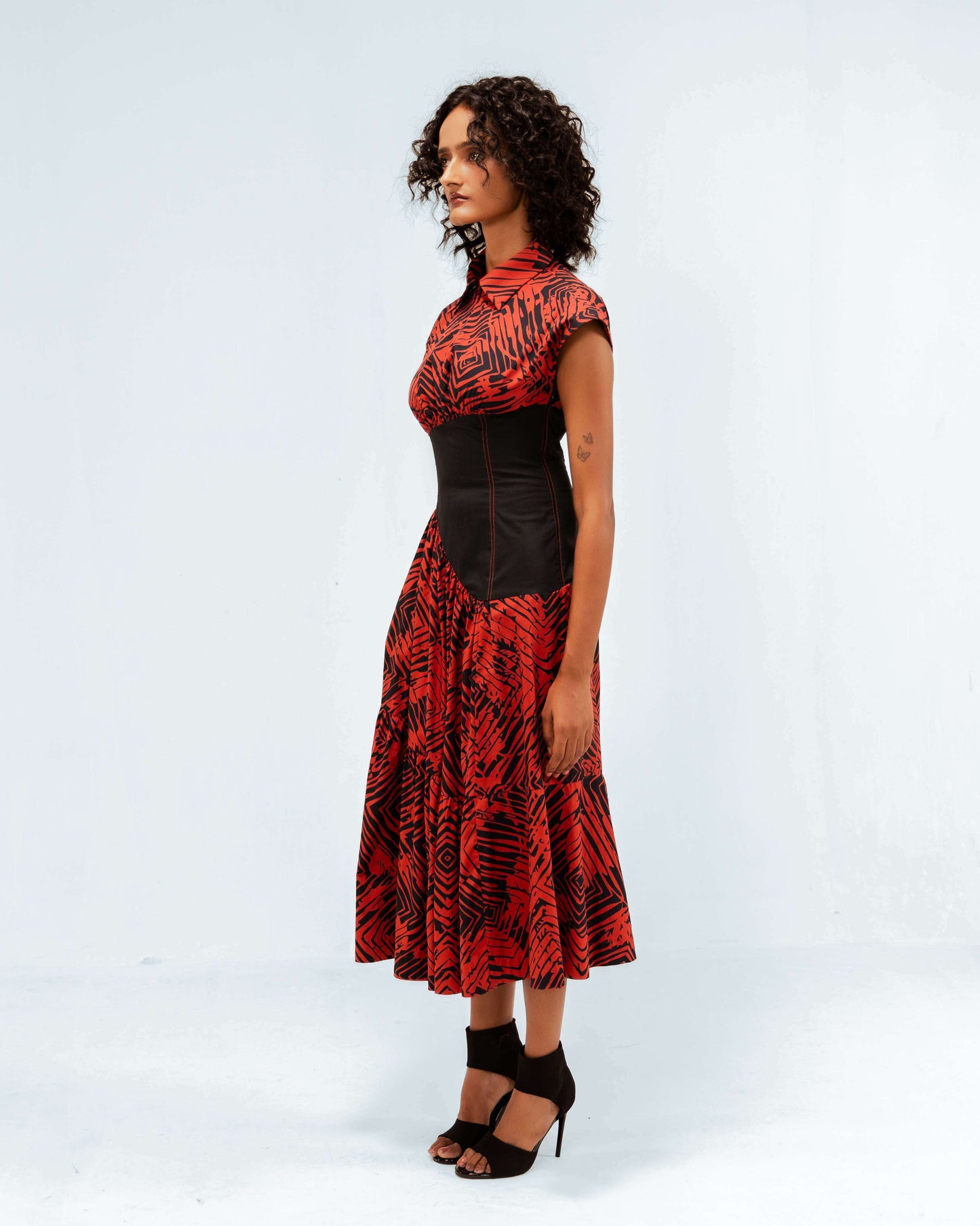 A model in profile wearing the Leh Studios Florix Print Basque Tiered Dress, an elegant piece from the Rhythm of time collection. This luxury upcycled fashion dress blends contemporary design with Indian traditions, featuring a collared neckline, cap sleeves, a striking red and black pattern, and a fitted black basque, perfect for eco-friendly women's apparel.