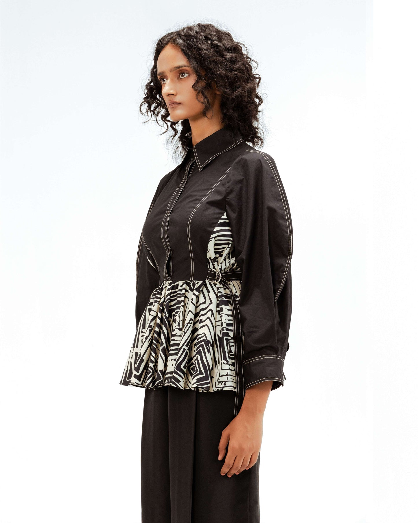 Model showcasing the Leh Studios Fable top from the Rhythm of Time collection, a black organic cotton shirt featuring prominent white contrast stitching and a gathered peplum waist with a bold black and white abstract print, highlighting sustainable Indian fashion and artisanal craftsmanship.