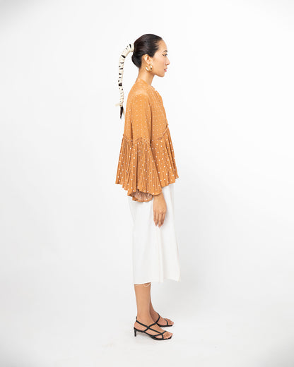 Woman wearing a mustard yellow top with ruffled sleeves and white pants on a white background from Leh Studios.