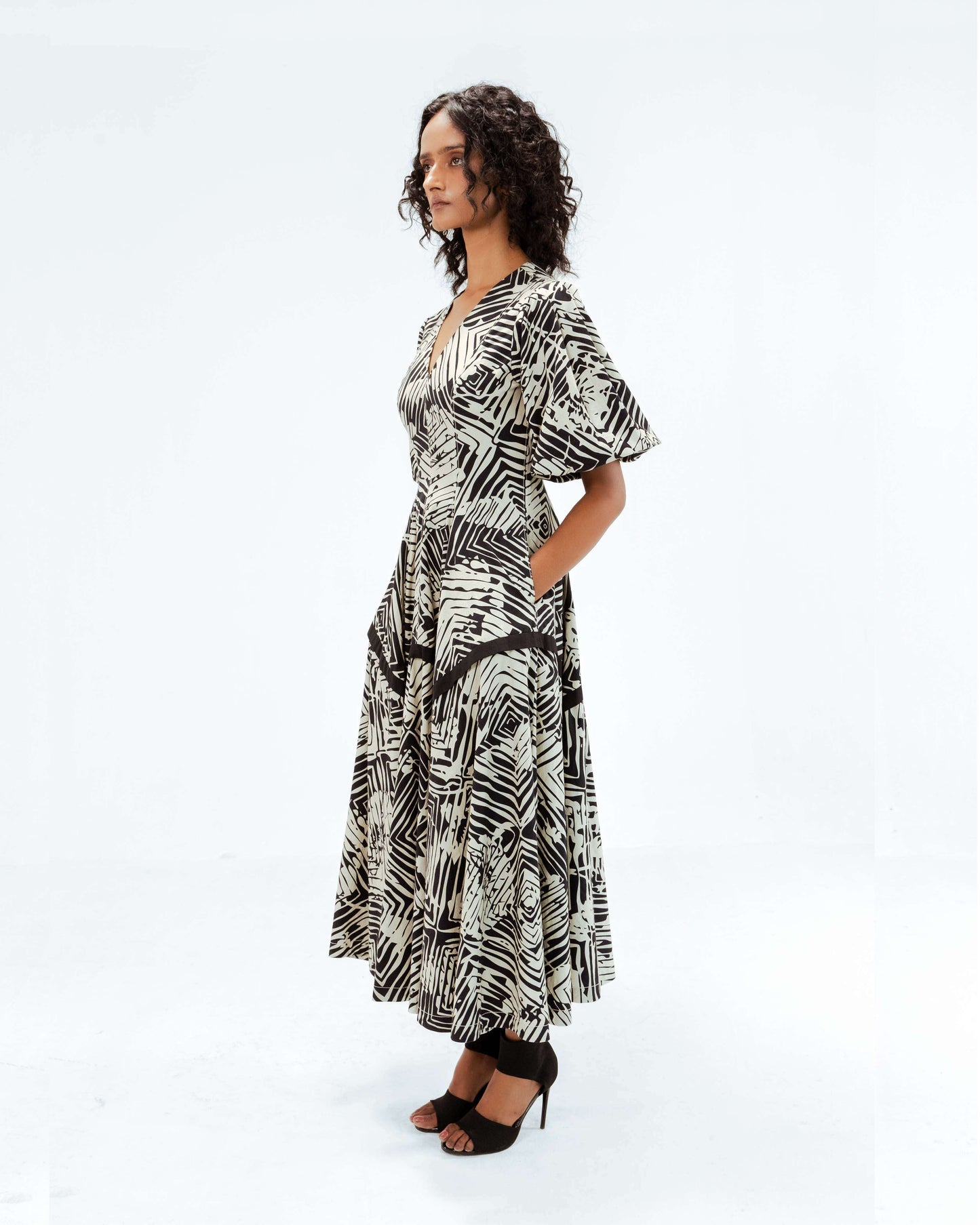 A model wears the Leh Studios Florix Print Thalia Dress, a luxurious upcycled fashion garment from the Rhythm of time collection. This black and cream patterned dress, with its defined waist and voluminous sleeves, embodies ethical fashion and artisanal craftsmanship, perfect for modern womens apparel.