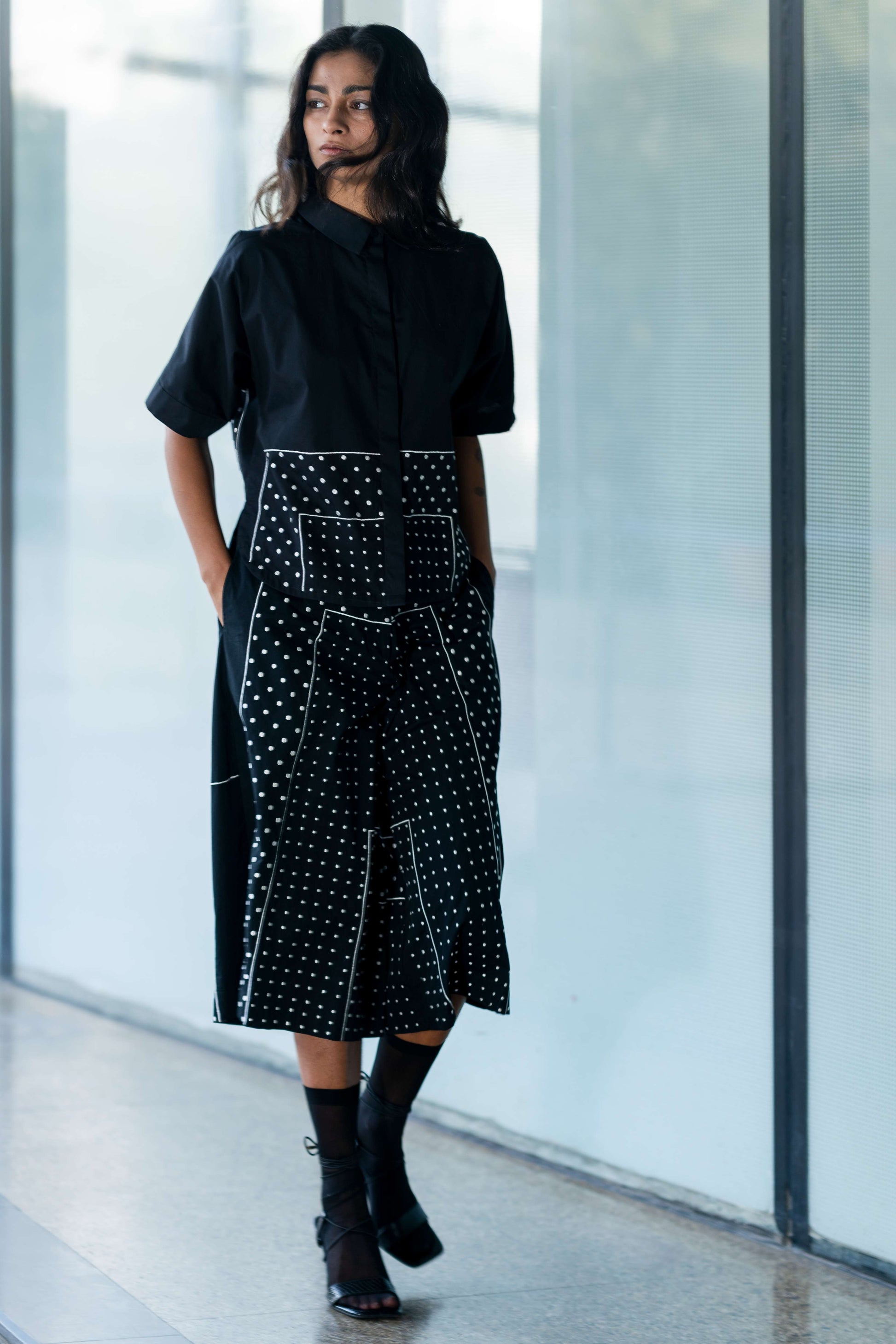 A person standing in a hallway wearing a black shirt with white polka dots embroidery and a black skirt from Leh Studios.