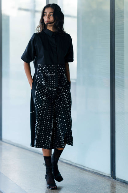 A person standing in a hallway wearing a black shirt with white polka dots embroidery and a black skirt from Leh Studios.