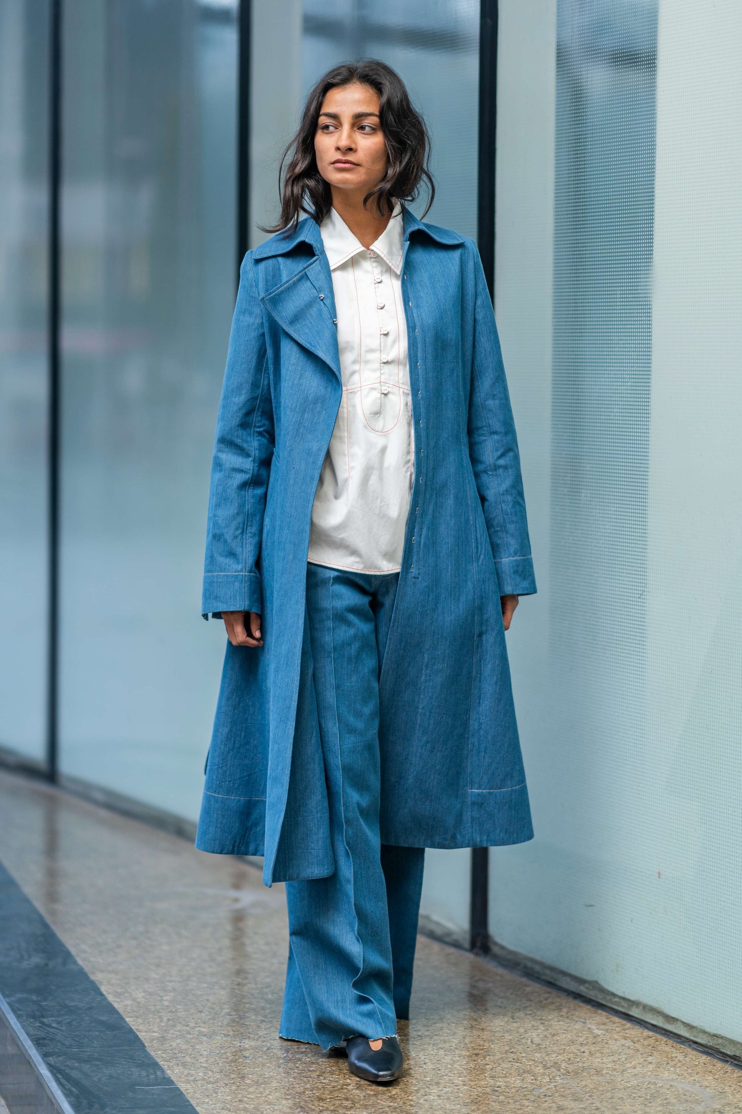 A woman wearing a blue long jacket with asymmetrical front opening and a tie up waist belt, paired with a white top and blue trousers, standing in a public space with reflective windows in the background from Leh Studios .