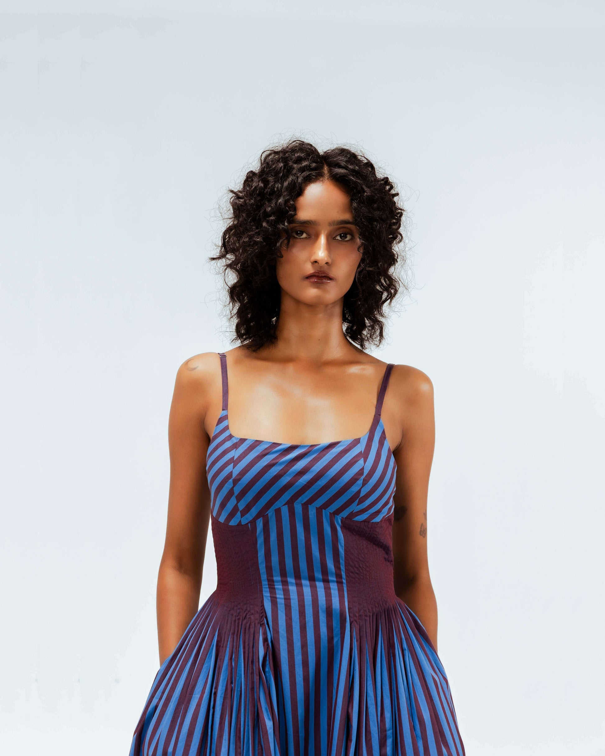 Model wearing the Leh Studios Meva Dress from the Rhythm of Time collection. This sustainable fashion piece features a blue and maroon striped design with a fitted bodice, thin straps, and a voluminous pleated skirt, showcasing eco-friendly design.