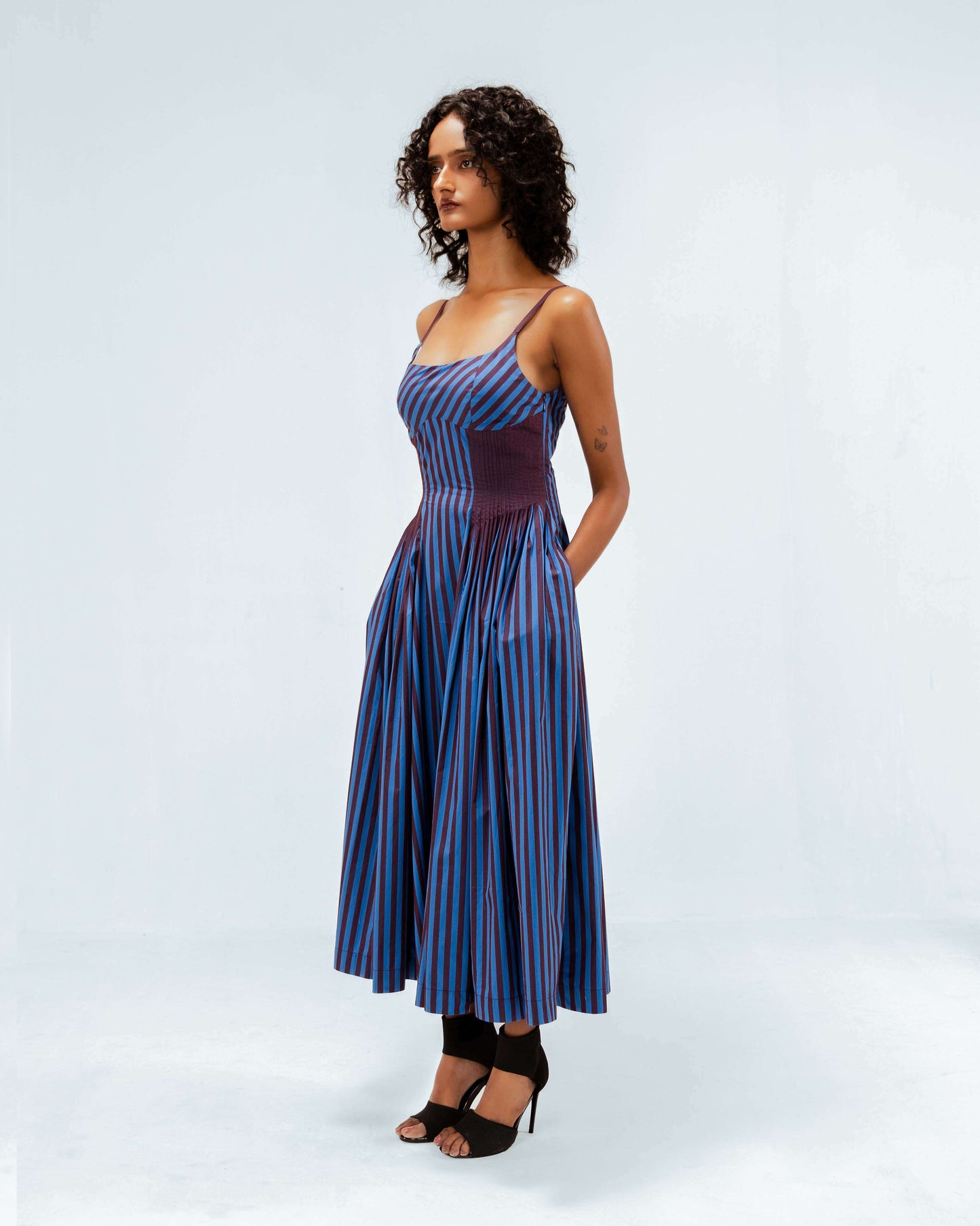 A model showcases the Leh Studios Meva Dress from the Rhythm of Time collection, a sustainable fashion midi-dress featuring elegant blue and maroon vertical stripes. This unique womens apparel piece boasts a fitted bodice, spaghetti straps, and a gracefully pleated skirt, embodying eco-friendly design and ethical fashion principles.