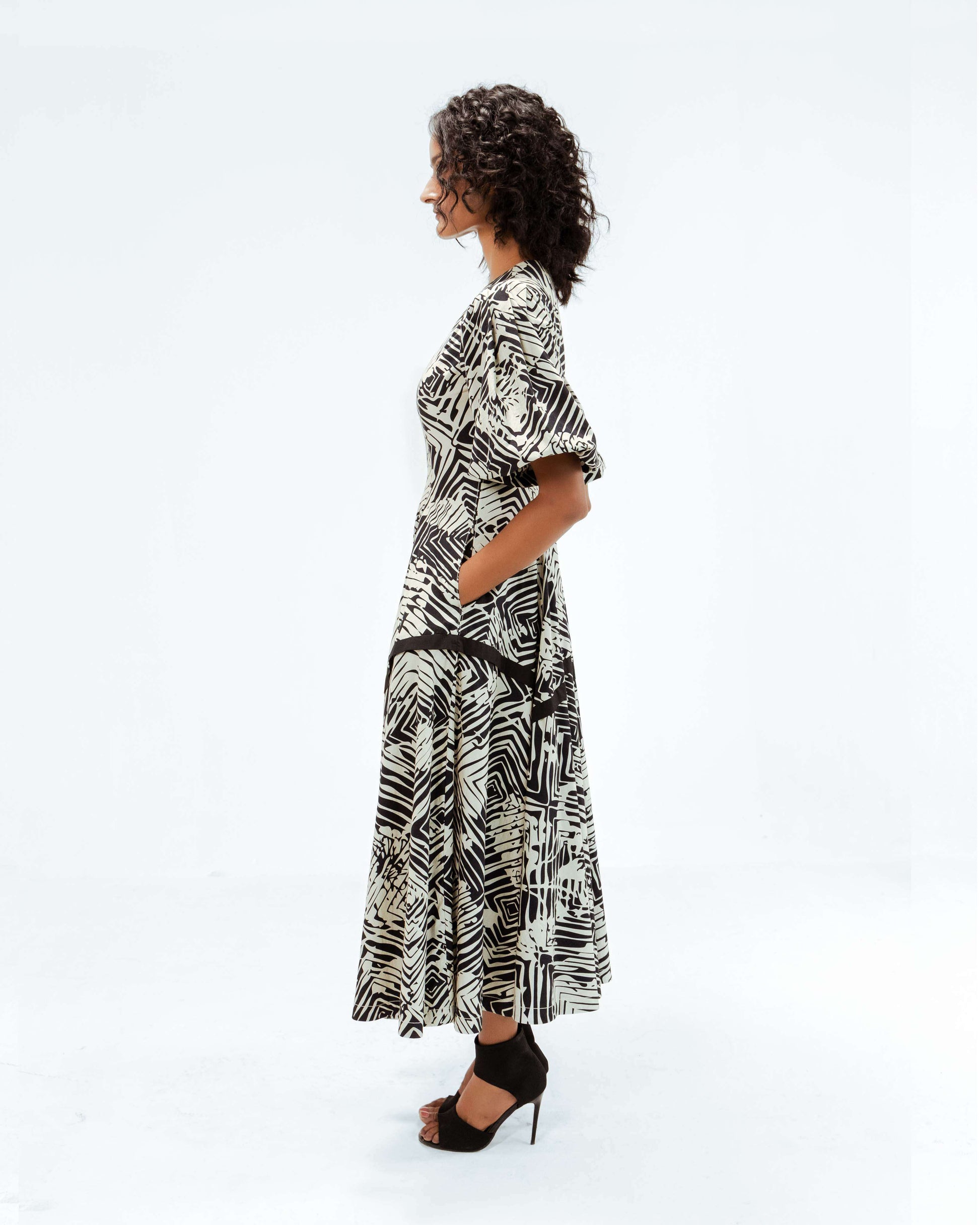 Leh Studios Florix Print Thalia Dress from the Rhythm of time collection, featuring an elegant black and off-white abstract pattern. This sustainable fashion piece offers contemporary designer wear for women, made with an eco-friendly design. The model stands in profile, showcasing the dress's sophisticated silhouette, puffed sleeves, and practical pockets.