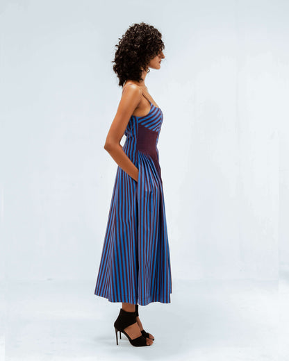 A model in profile showcases the Leh Studios Meva Dress from the Rhythm of time collection, a blue and maroon striped midi dress with spaghetti straps and convenient side pockets. This piece embodies sustainable fashion and handcrafted apparel, perfect for womens apparel seeking ethical designer wear.