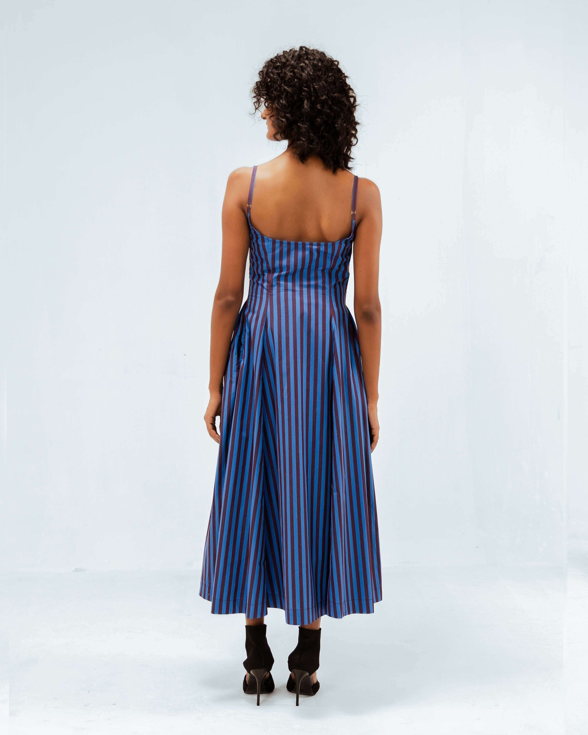 Model showcasing the Leh Studios Meva Dress from the Rhythm of time collection, a blue and maroon striped midi dress with delicate spaghetti straps, viewed from the back. This sustainable fashion piece is crafted with an emphasis on organic cotton, reflecting designer wear for sophisticated womens apparel.