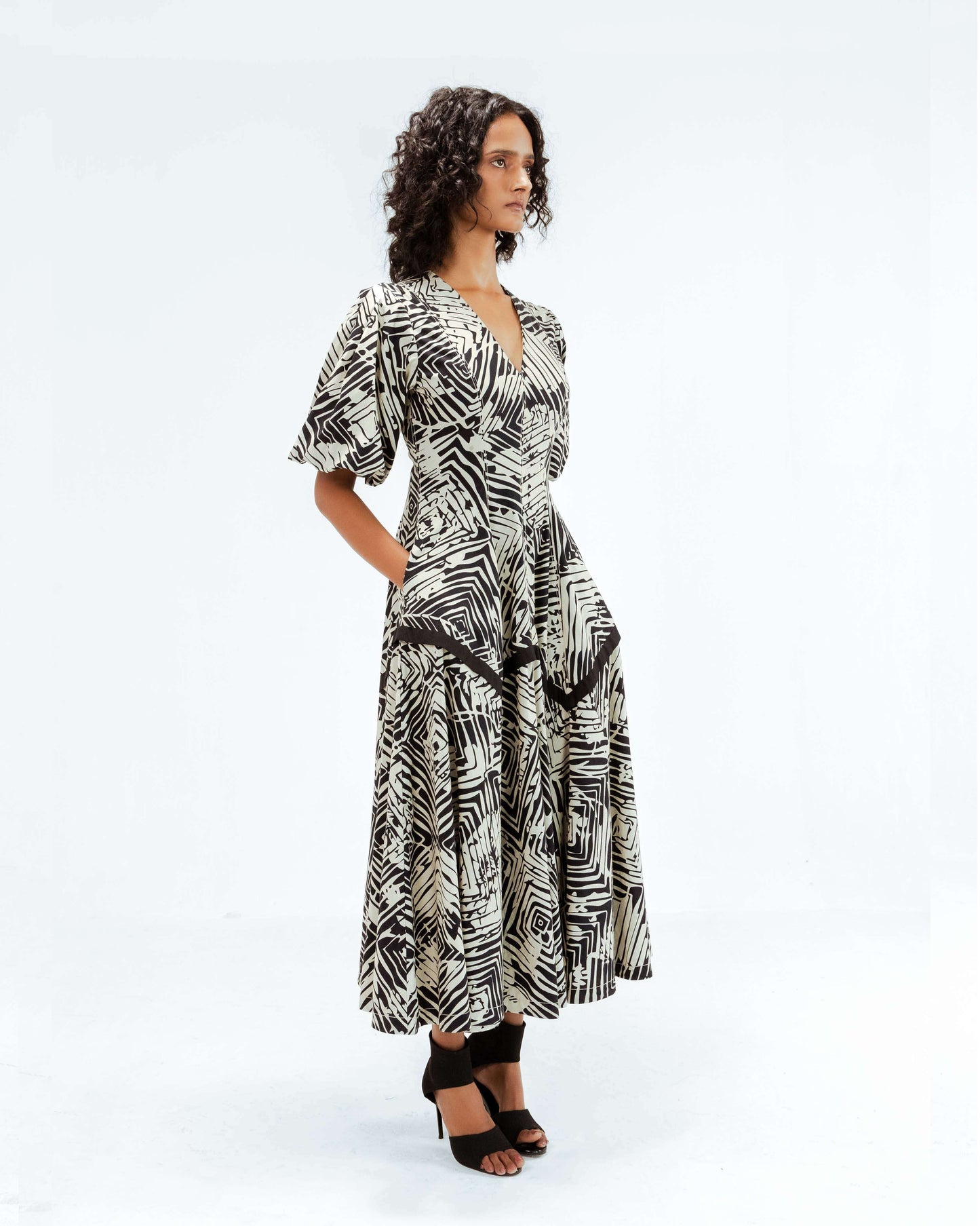 A model showcases the Leh Studios Florix Print Thalia Dress from the Rhythm of time collection, a stunning black and white abstract-patterned midi dress made from organic cotton. This sustainable fashion piece embodies ethical designer wear, featuring a flattering V-neck, elegant puff sleeves, and a full, flowing skirt, perfect for contemporary womens apparel. The model stands with hands casually in pockets.