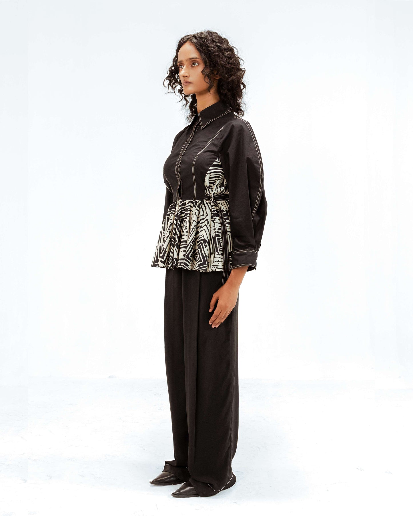 Full-body side profile of a model wearing the Leh Studios Fable top, a black button-up blouse with prominent white stitching and an upcycled black and white abstract print panel around the waist, part of the eco-friendly Rhythm of time collection. This luxury upcycled fashion piece highlights unique designer wear.