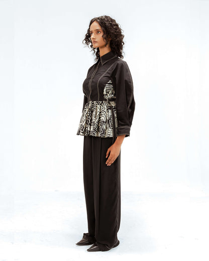 Full-body side profile of a model wearing the Leh Studios Fable top, a black button-up blouse with prominent white stitching and an upcycled black and white abstract print panel around the waist, part of the eco-friendly Rhythm of time collection. This luxury upcycled fashion piece highlights unique designer wear.