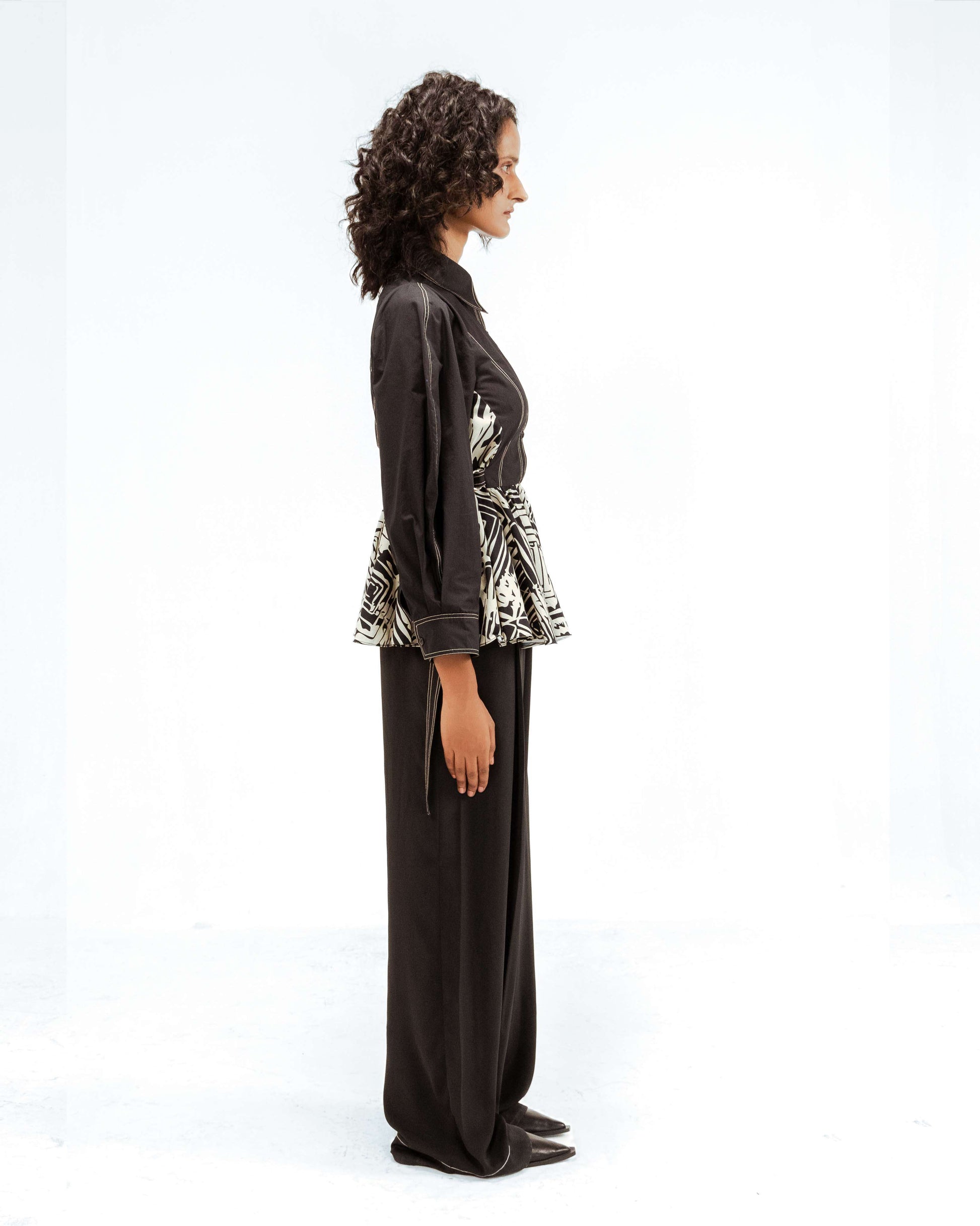A model in profile showcases the Leh Studios Fable top from the Rhythm of time collection. This sustainable Indian fashion piece features a dark base with a striking black and white geometric pattern around the peplum and intricate stitching details, reflecting ethical craftsmanship and designer wear.