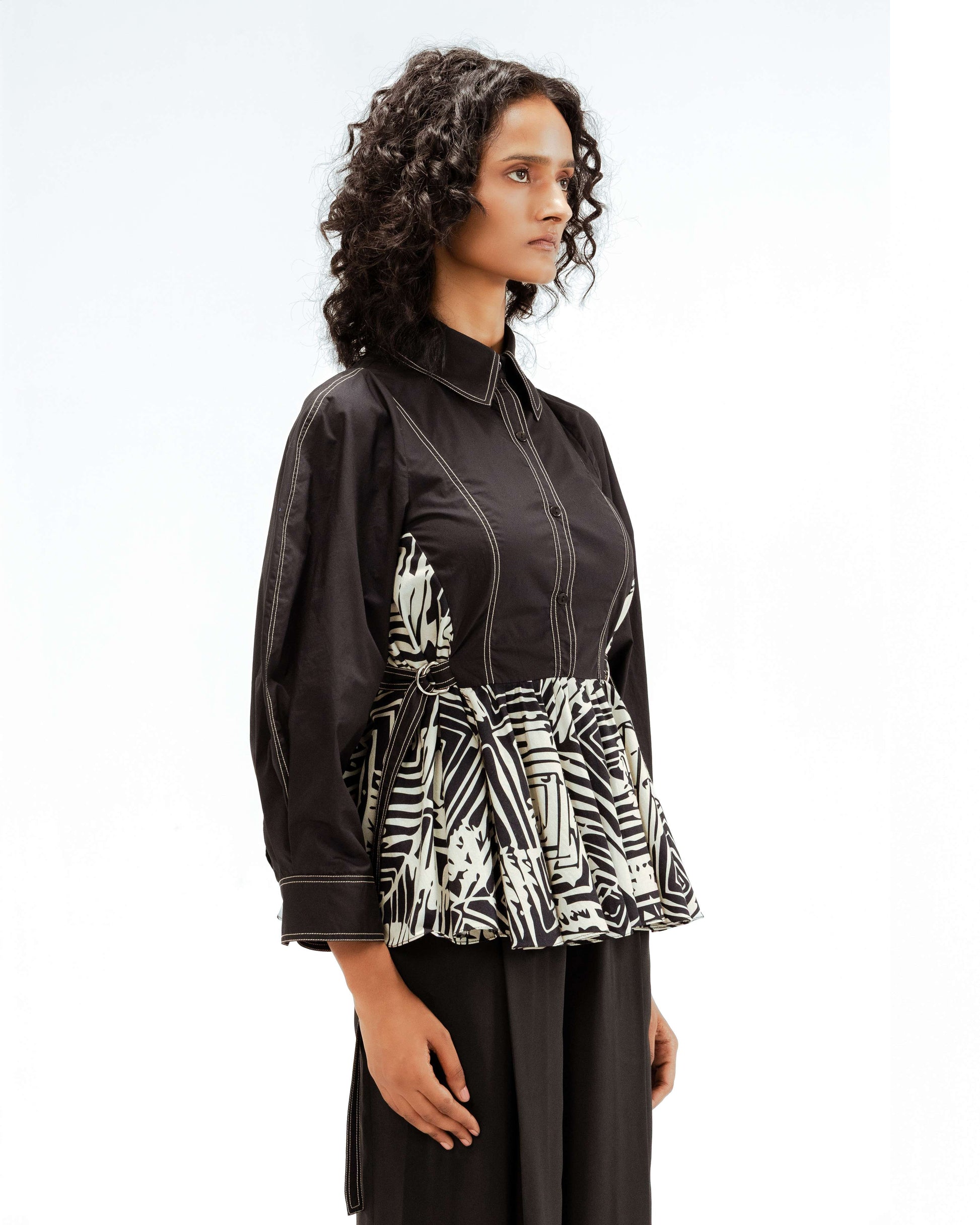 A model showcasing the Leh Studios Fable top from the Rhythm of time collection, featuring a black organic cotton shirt with crisp white contrast stitching and a striking black and white geometric print peplum waist. This sustainable fashion piece embodies eco-friendly designer wear for women.