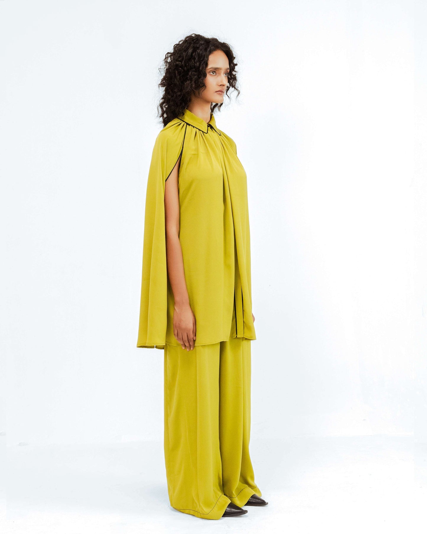 A model showcasing the Leh Studios Cherice Crepe Cordset from the Rhythm of time collection, featuring a vibrant chartreuse cape-style top with black trim and wide-leg trousers. This sustainable fashion designer wear embodies eco-friendly design and artisanal craftsmanship for contemporary womens apparel.